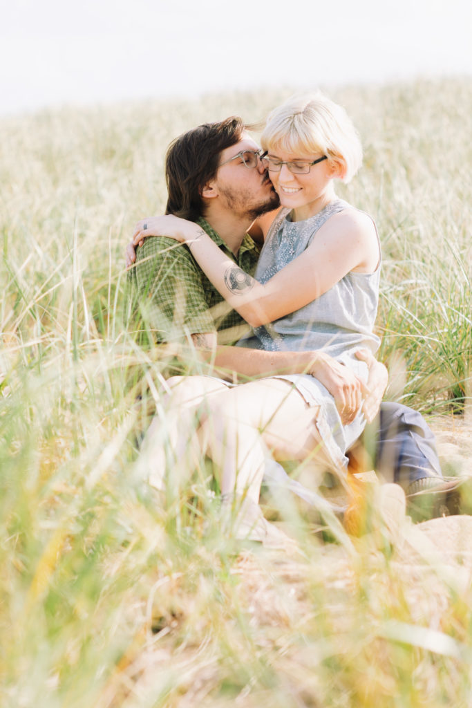 Woman sits on mans lap and man kisses her cheek in the middle of a field on a beach