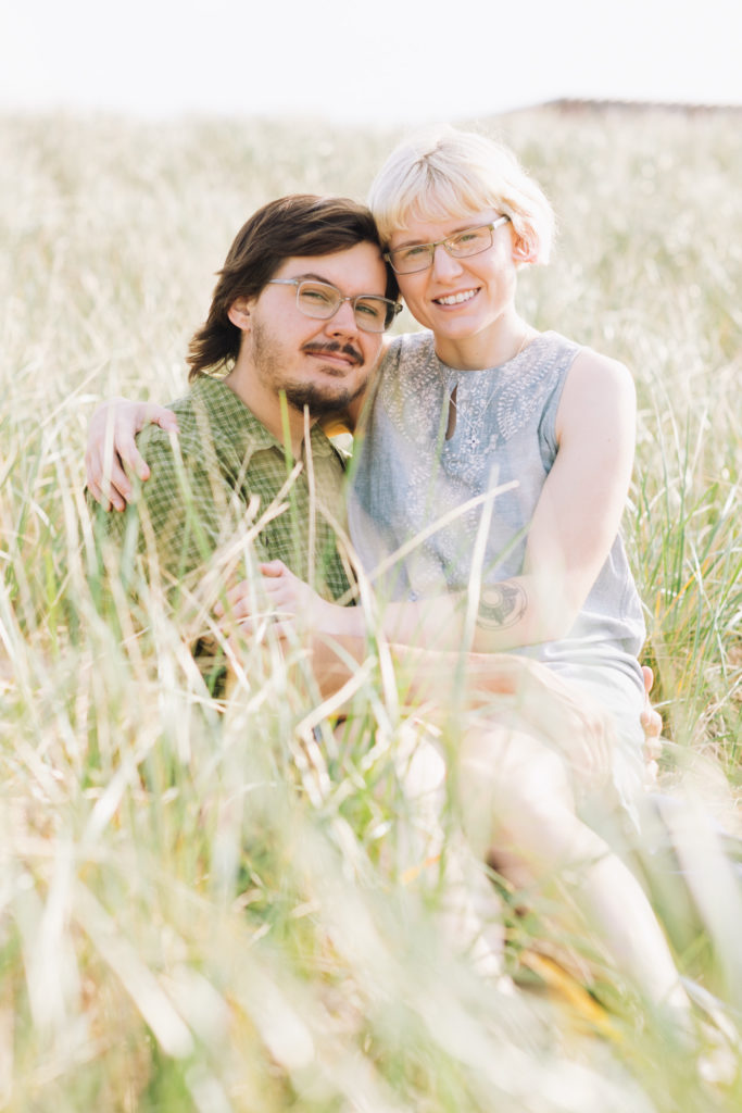 Man and woman look at camera and smile while sitting on each others laps on the beach grass